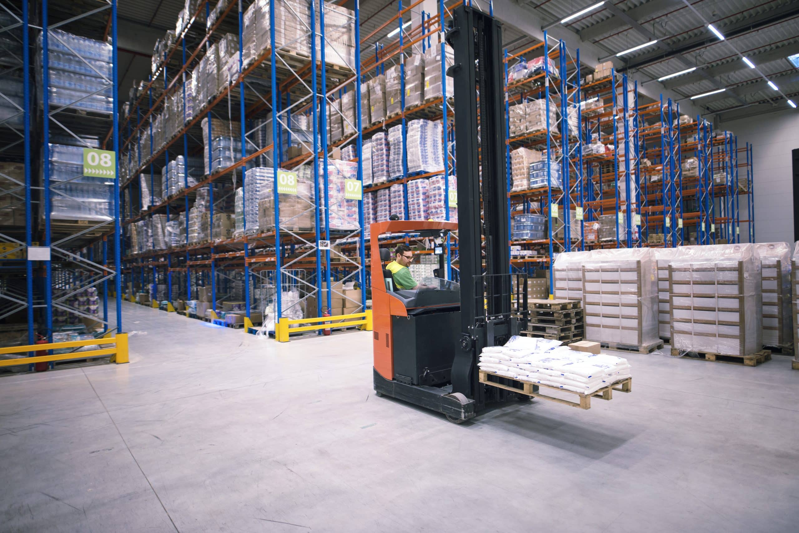 Worker operating forklift machine and relocating goods in large warehouse centre.